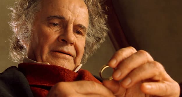 A close-up photograph of an older man with curly gray hair, wearing a dark coat and a red scarf, holding a small golden ring between his fingers. The image is a still from the film 'The Lord of the Rings: The Fellowship of the Ring,' featuring the character Bilbo Baggins. The man is looking down at the ring with a contemplative expression.