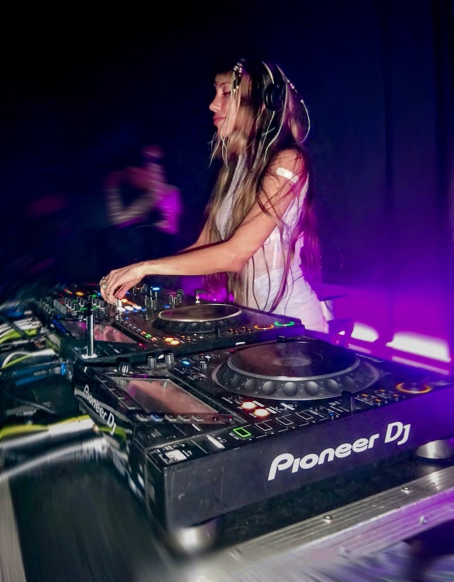 A woman with long hair and headphones is shown in profile, operating a Pioneer DJ mixer and turntable setup in a dark room illuminated by purple light.