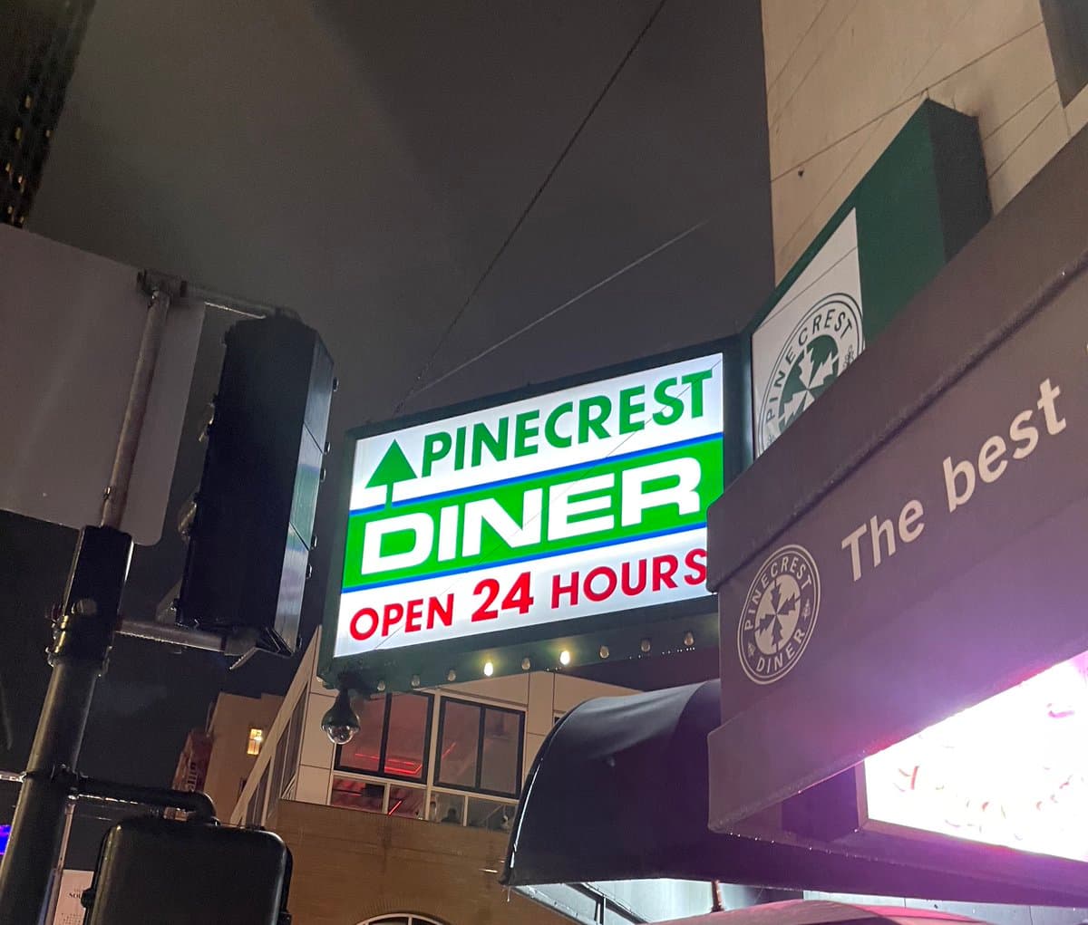 A nighttime street-level photograph looking up at the illuminated signage for Pinecrest Diner. The main sign displays the restaurant's name and hours, while a secondary sign on the right features the slogan 'The best'.
