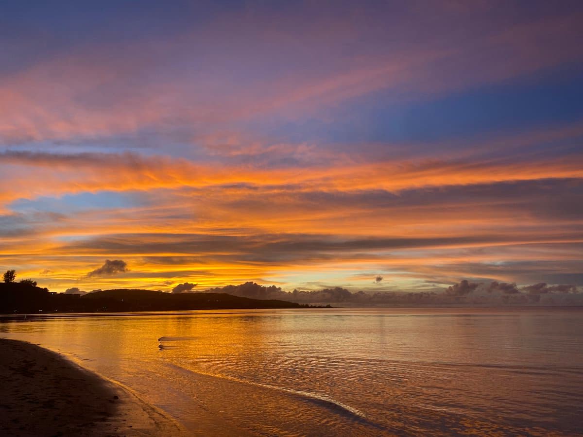 A scenic photograph of a sunset over a calm body of water, featuring a sky filled with orange and purple clouds reflecting on the surface. A dark, silhouetted coastline is visible in the distance, with a sandy beach in the foreground.