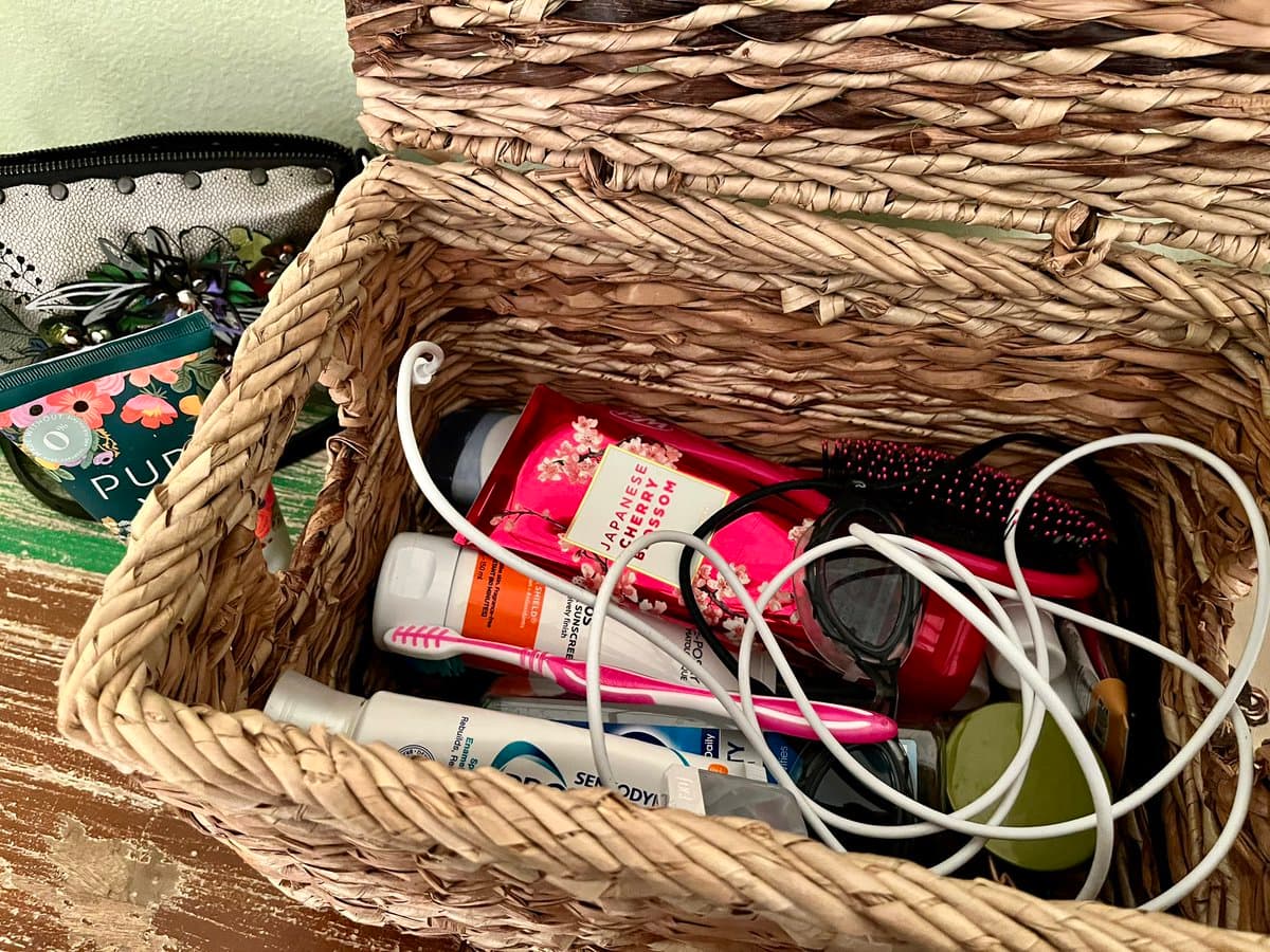 A top-down view of a woven wicker basket filled with various toiletries, including a pink tube of Japanese Cherry Blossom lotion, a white bottle of sunscreen, a pink toothbrush, and a tangled white charging cable.