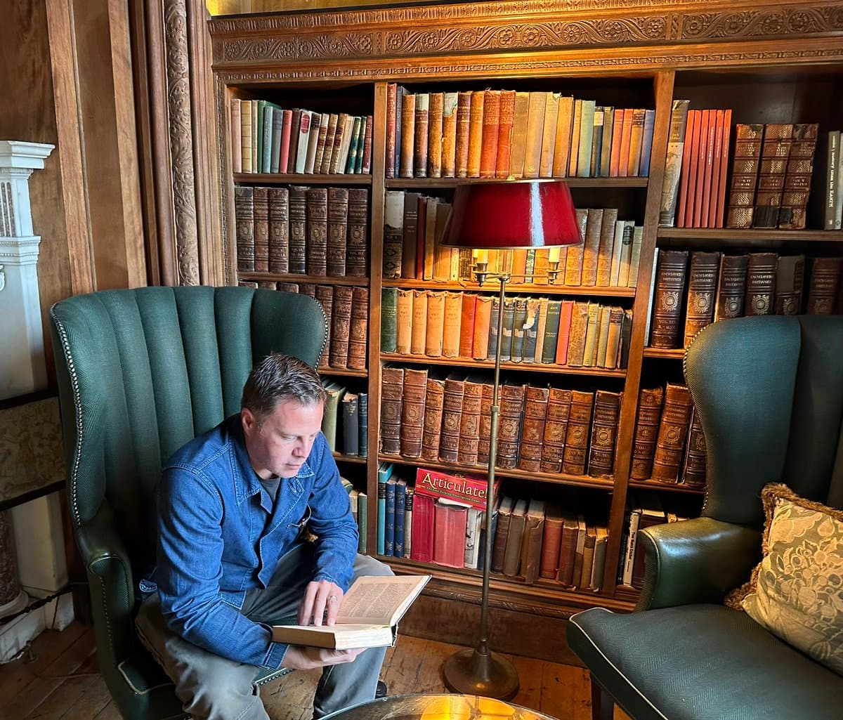 A man in a blue denim jacket sits in a green wingback chair, reading a book in a library with floor-to-ceiling wooden bookshelves filled with antique volumes. A red-shaded floor lamp stands between the chairs, and a second green armchair is visible on the right.