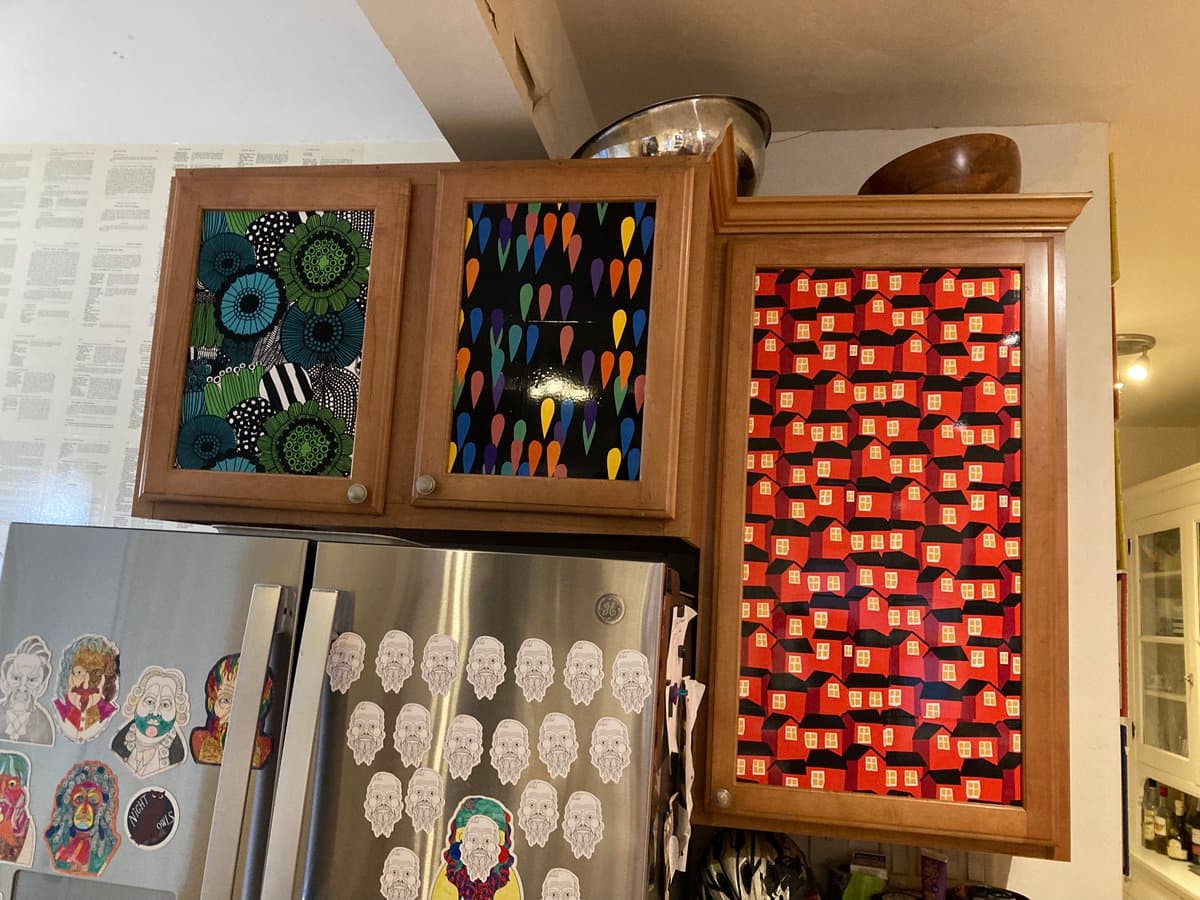 A photograph of a kitchen featuring wooden cabinets decorated with colorful, patterned Marimekko fabric panels. A stainless steel refrigerator is visible in the foreground, covered with various stickers and magnets.