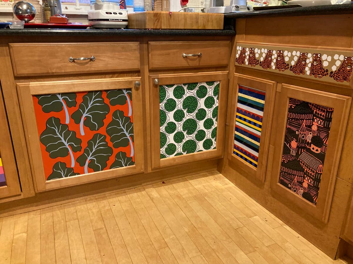 A photograph of a kitchen corner featuring light wood cabinets decorated with colorful, patterned Marimekko fabric panels on the doors and a tiger-print border. The countertop holds various kitchen items, including a KitchenAid appliance and a wooden cutting board.