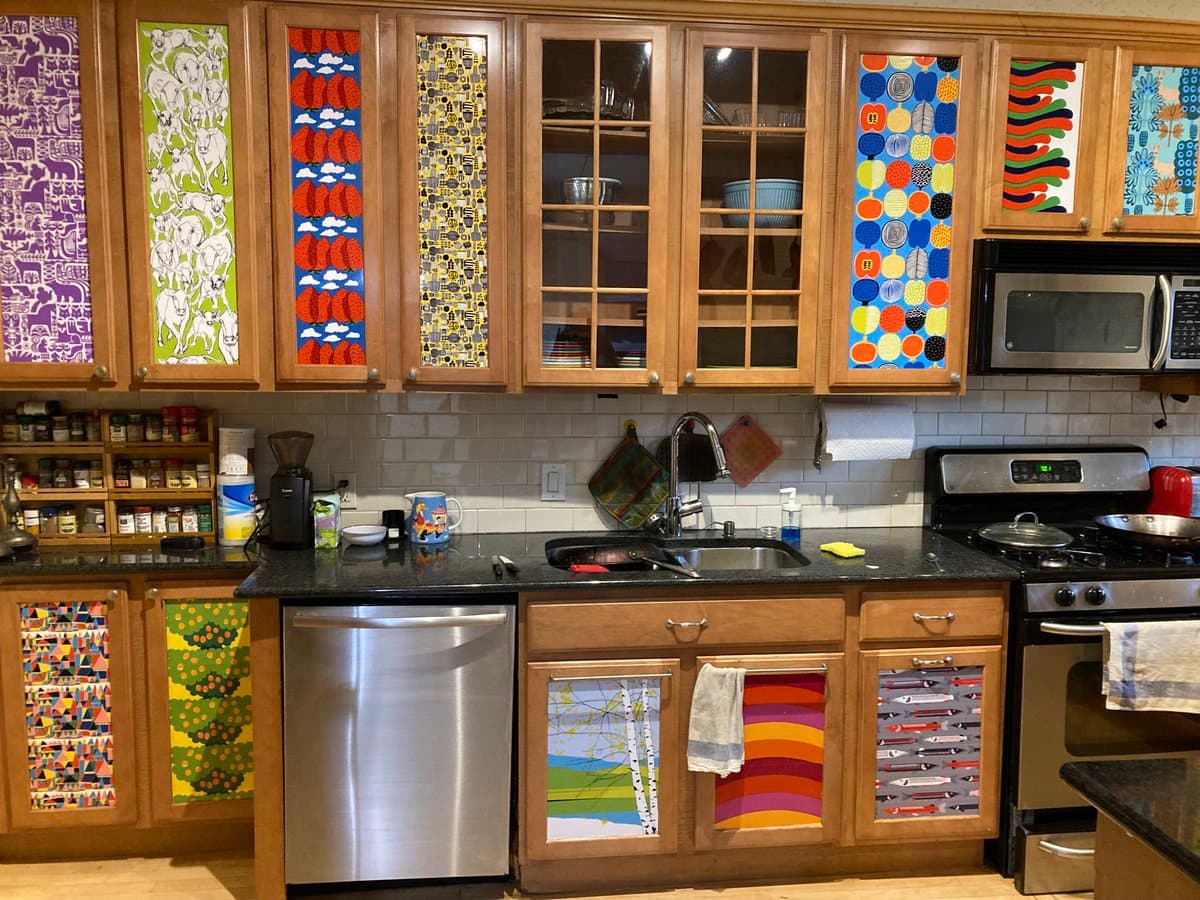 A photograph of a kitchen featuring wooden cabinets with doors decorated in various colorful, patterned Marimekko fabric designs. The kitchen includes a stainless steel dishwasher, a black countertop, and a white subway tile backsplash.
