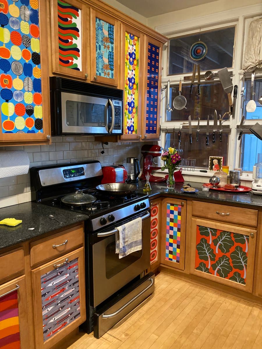 A photograph of a kitchen corner featuring wooden cabinets with doors decorated in various colorful, patterned Marimekko fabrics. The kitchen includes a stainless steel stove and microwave, a black countertop, and a window with hanging cooking utensils.