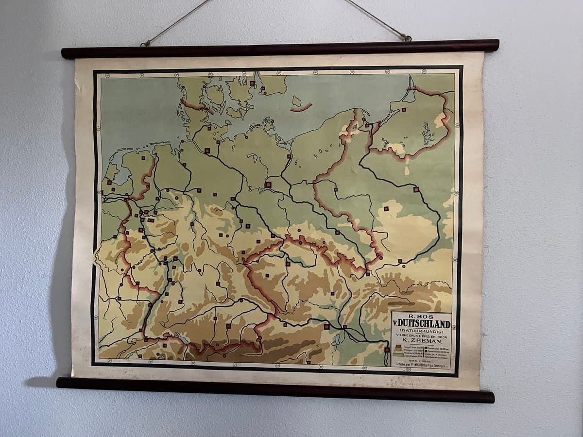 A vintage-style topographic map of Germany is displayed on a wall between two wooden dowels. The map, titled 'R. BOS V. DUITSLAND (NATUURKUNDIG) VERDEELING DOOR K. ZEEMAN', features color-coded elevation zones and various geographic markers.