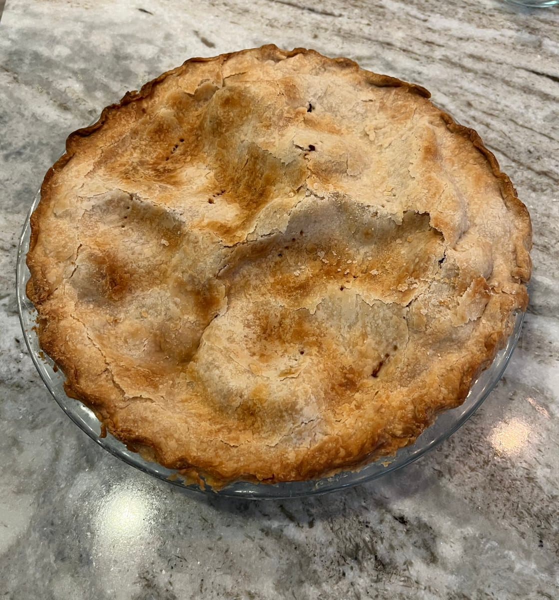 A freshly baked, golden-brown pie with a fluted crust sits in a clear glass dish on a gray marble countertop. The top crust is uneven and rustic, with visible cracks and patches of flour dusting.