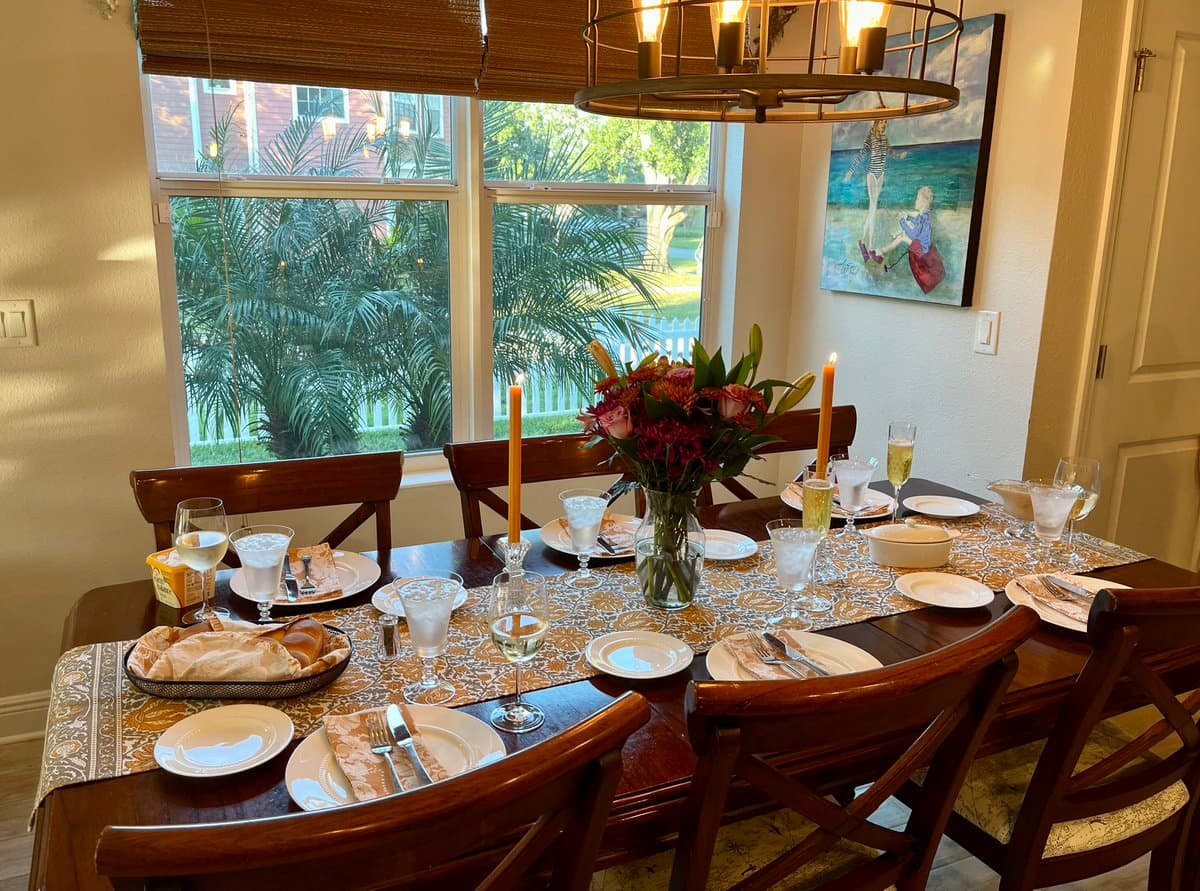 A dining table is set for a meal with white plates, silverware, wine glasses, and water glasses, featuring a floral centerpiece and lit candles. The room has large windows looking out onto a garden with palm trees, and a painting hangs on the wall.