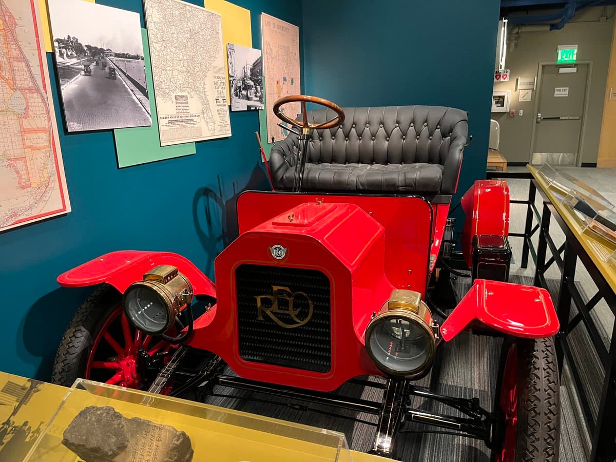 A red vintage automobile with a black leather bench seat and brass headlights is displayed in a museum exhibit. The car is positioned in front of a blue wall decorated with historical maps and photographs, with a glass display case visible in the foreground.