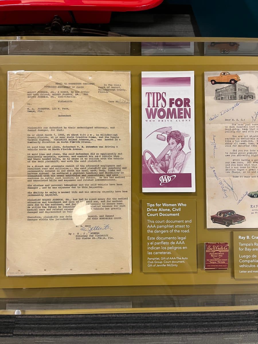 A museum display case featuring historical documents related to a 1966 car accident and a 1970s AAA pamphlet titled 'Tips for Women Who Drive Alone.' The exhibit includes a legal statement of claim, the pamphlet itself, and a letter from Ray B. Craddock, accompanied by informational placards in English and Spanish.