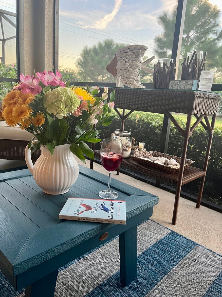 A glass of red sangria and a copy of the book 'This Is How You Lose the Time War' sit on a blue wooden table next to a white pitcher of colorful flowers. In the background, a wicker console table holds a driftwood sculpture and a sign, all set on a screened-in porch with a view of trees.