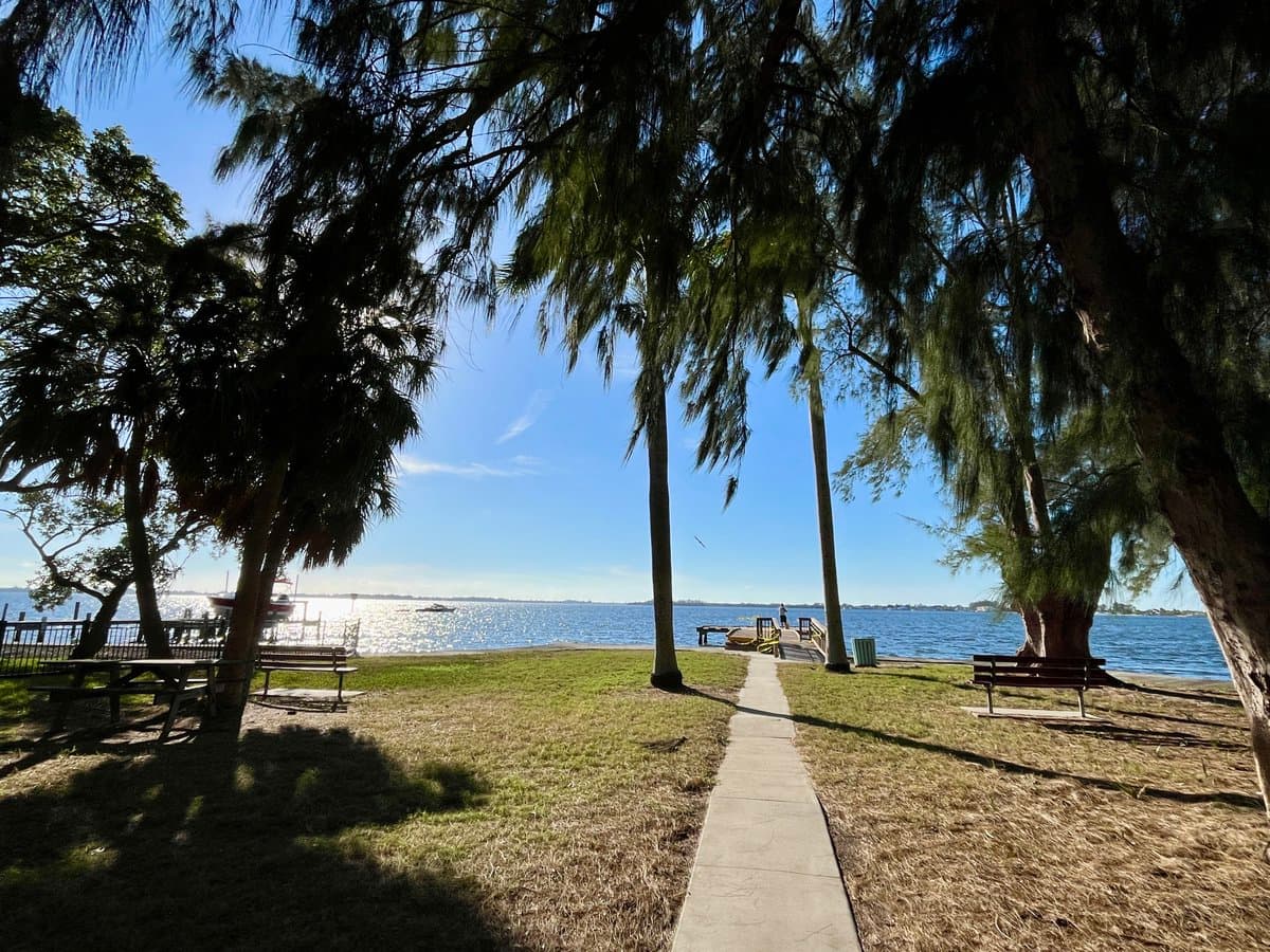 A scenic outdoor photograph of a park area overlooking a body of water on a sunny day. The foreground features a paved walking path, grassy areas, and several benches and picnic tables shaded by tall trees, with a boat visible on the water in the distance.
