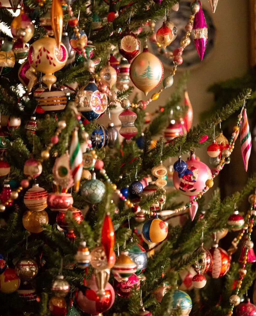 A close-up photograph of a Christmas tree densely decorated with a variety of colorful, vintage-style glass ornaments and beaded garlands.