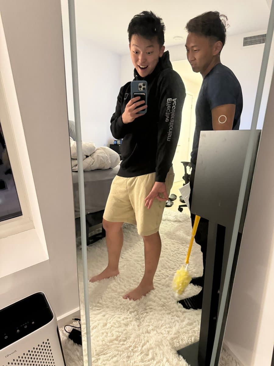 A mirror selfie of a young man in a black hoodie and beige shorts, smiling while holding a smartphone, with another man standing behind him in a bedroom. The man in the back is holding a yellow duster. The room features a white shag rug, a bed, and a white air purifier.