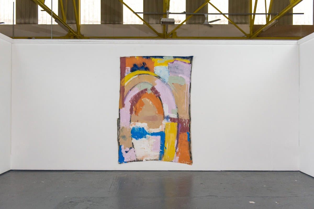 A wide shot of a colorful abstract painting hanging on a white gallery wall, featuring a prominent arch shape in orange and purple tones. The room has a polished concrete floor and an industrial ceiling with yellow metal beams.