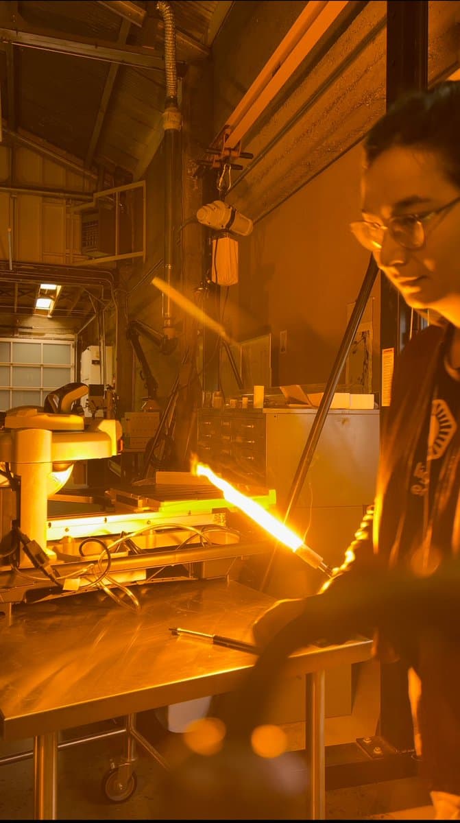 A person wearing safety glasses works in a workshop, holding a long glass rod that is glowing bright orange from intense heat. The scene is illuminated by the warm light of the heated glass and industrial equipment in the background.