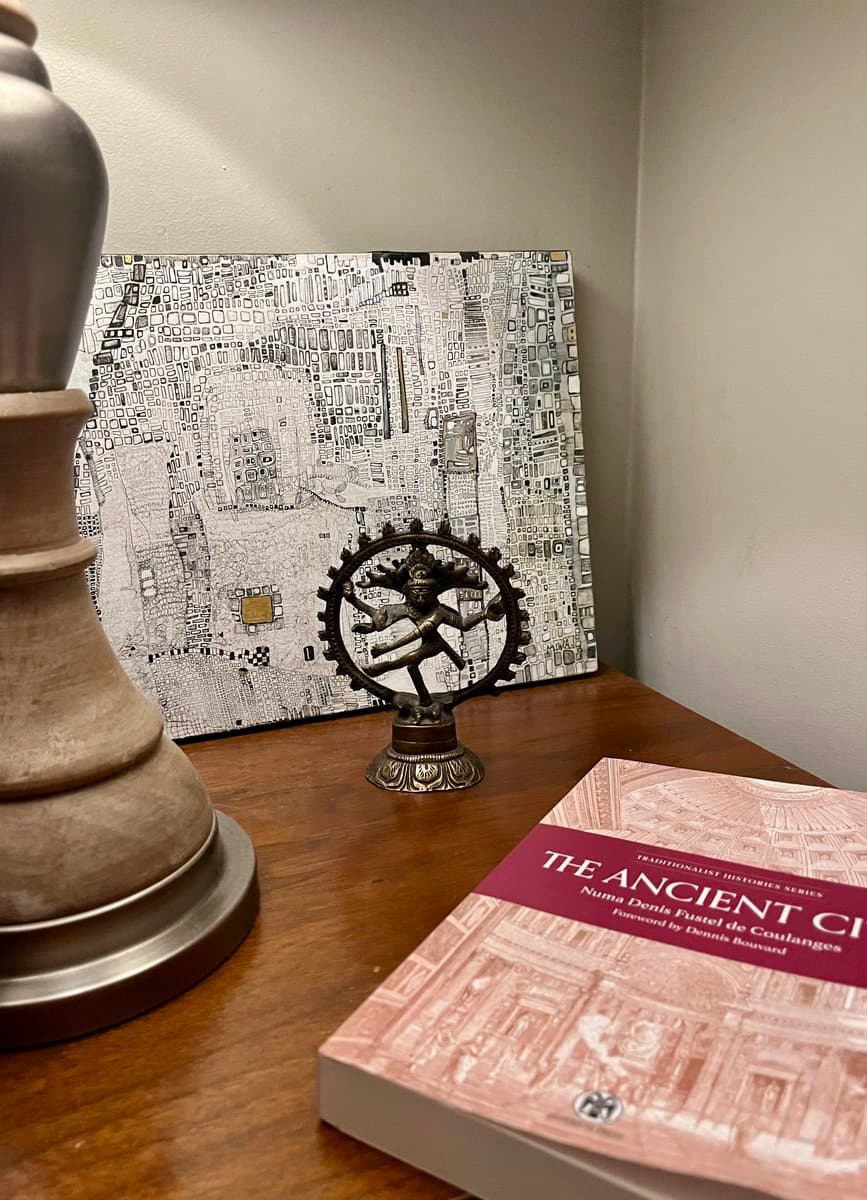A photograph of a wooden desk displaying a small bronze Nataraja statue, a large abstract mosaic canvas, and a book titled 'The Ancient City.'