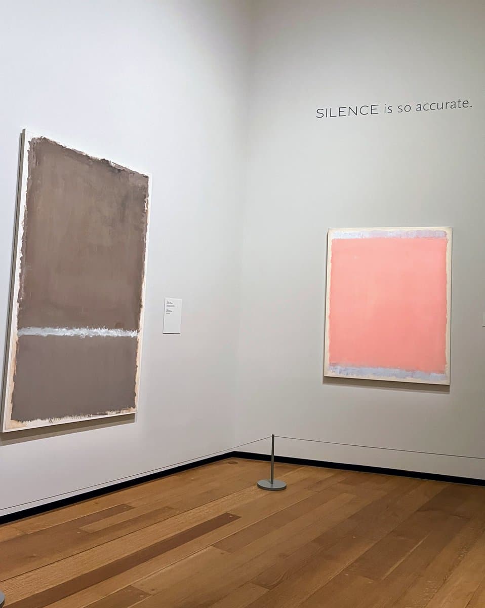 A photo of a gallery corner with two abstract paintings on white walls and a wooden floor. The text "SILENCE is so accurate." is projected onto the right wall above a pink and blue painting.