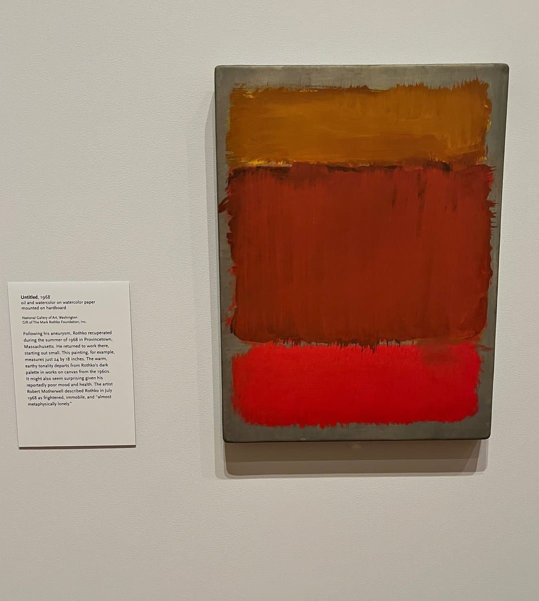 A photograph of a Mark Rothko abstract painting featuring horizontal bands of orange, red, and dark red on a gray background, displayed on a gallery wall next to an informational placard. The placard identifies the work as 'Untitled, 1968' and provides details about its creation and context.