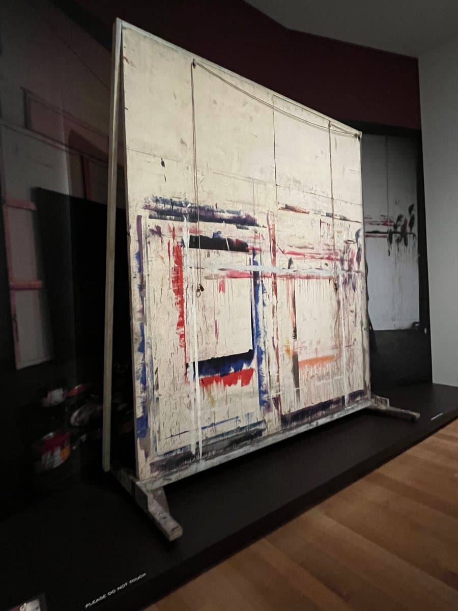 A large, abstract painting on a wooden easel is displayed in a gallery, featuring a white canvas with vertical drips and patches of red, blue, and black paint. The artwork rests on a black platform in a dimly lit room with a wooden floor.