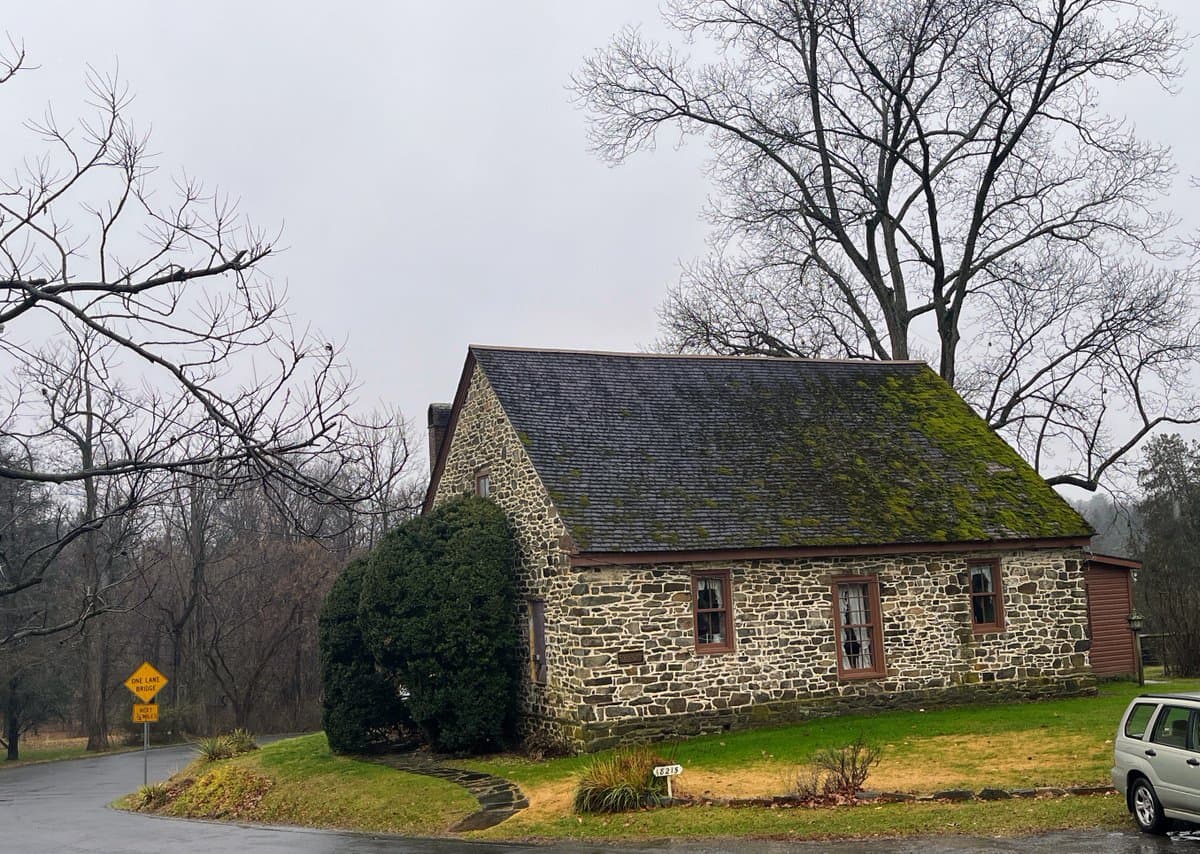 A stone house with a moss-covered roof sits on a grassy corner next to a wet, paved road on an overcast day. Bare trees frame the scene, and a silver SUV is partially visible on the right.