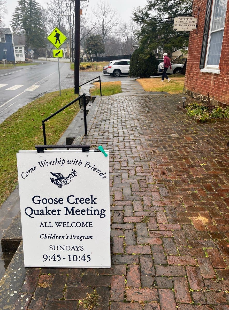 A photograph of a wet, brick-paved walkway leading to a red brick building on a rainy day. A white sign in the foreground advertises a Quaker meeting, while a person in a red jacket walks in the background near a parked white SUV.
