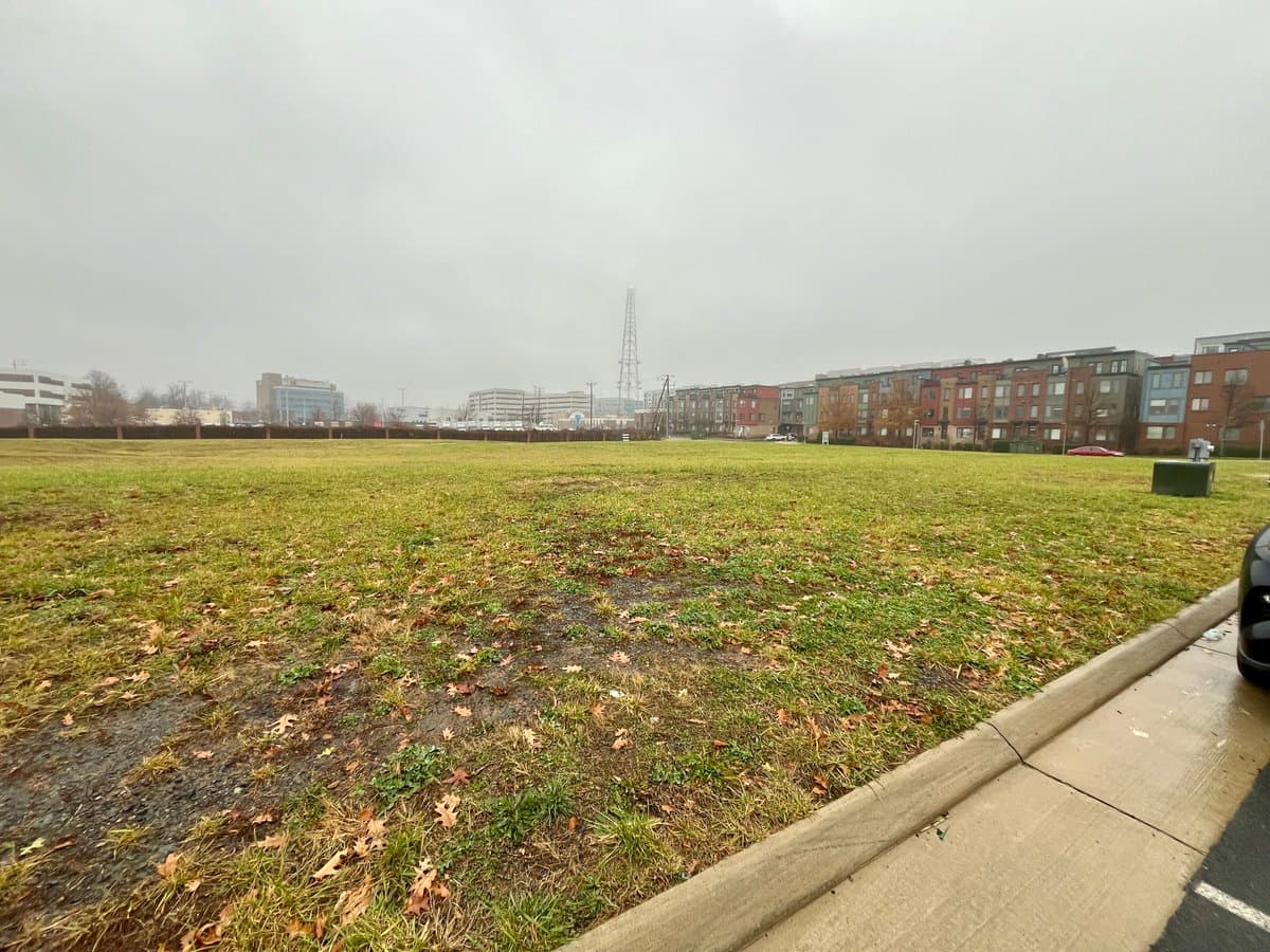 A wide shot of an empty, grassy field on an overcast day, bordered by a sidewalk and a row of multi-story residential buildings in the background.