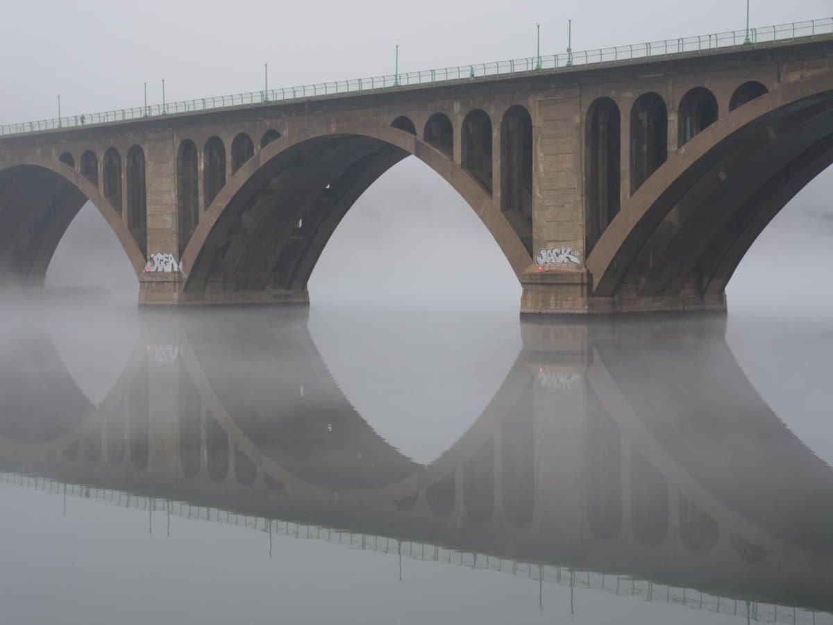 A multi-arched stone bridge spans a calm river, partially obscured by a thick layer of low-lying fog that creates a clear reflection on the water's surface.