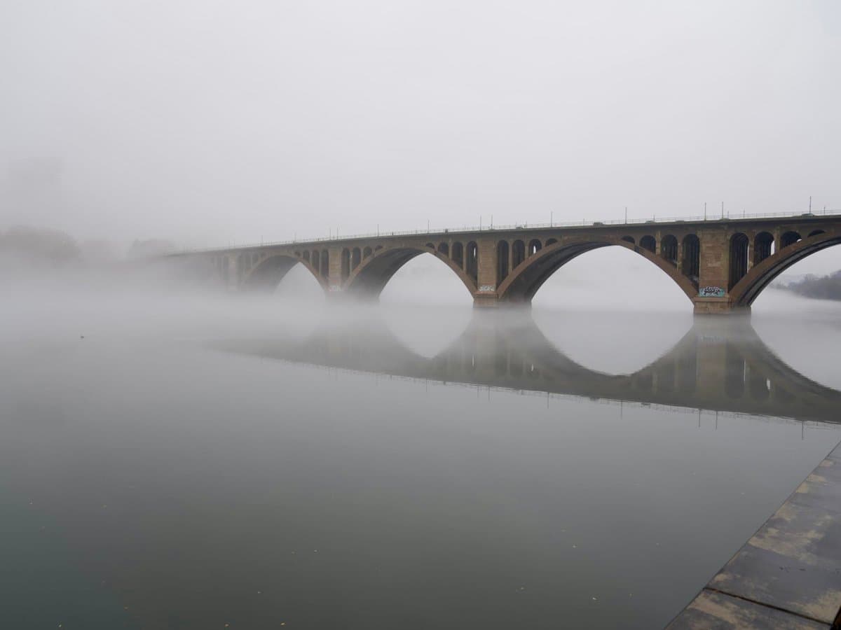 A multi-arched stone bridge spans a wide river, partially obscured by a thick layer of low-lying fog hovering over the water's surface.