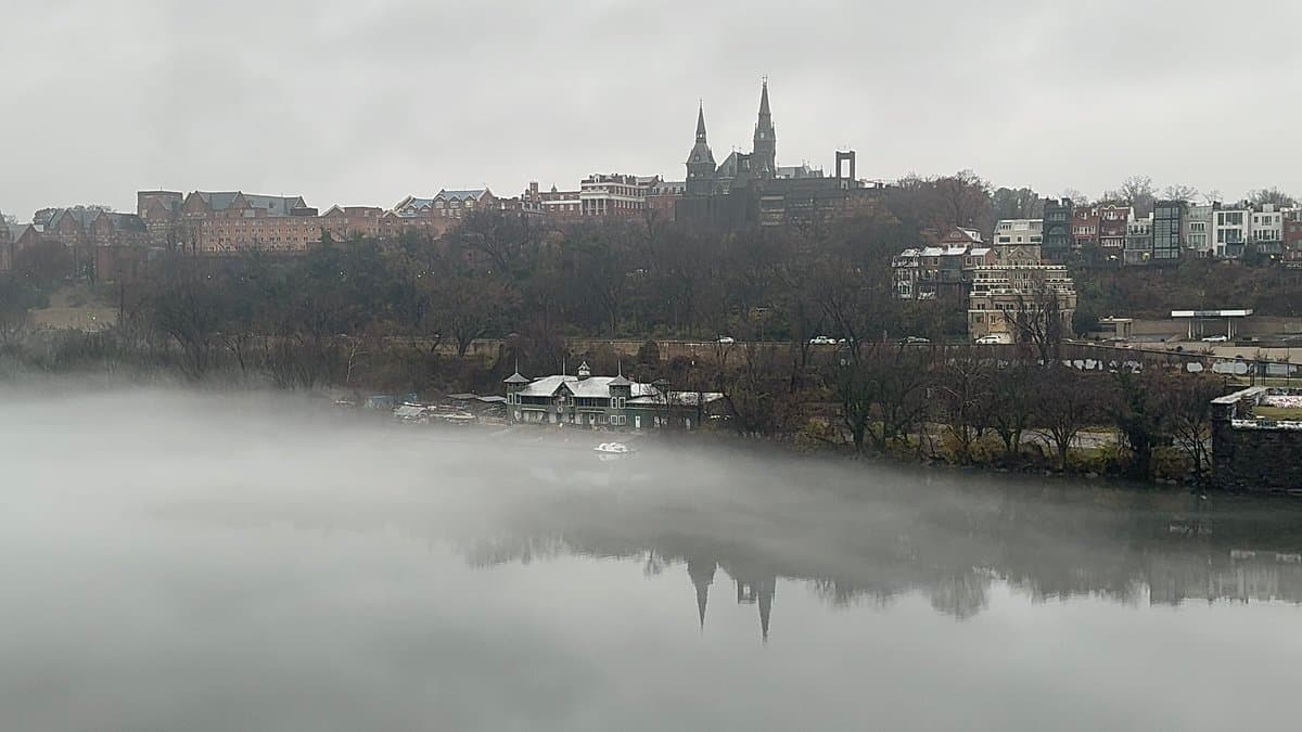A wide landscape photograph showing a city skyline on a hill across a river, partially obscured by a thick layer of low-lying fog hovering over the water. The scene includes a prominent church with twin spires and a small white building on the riverbank, all reflected in the calm water below.