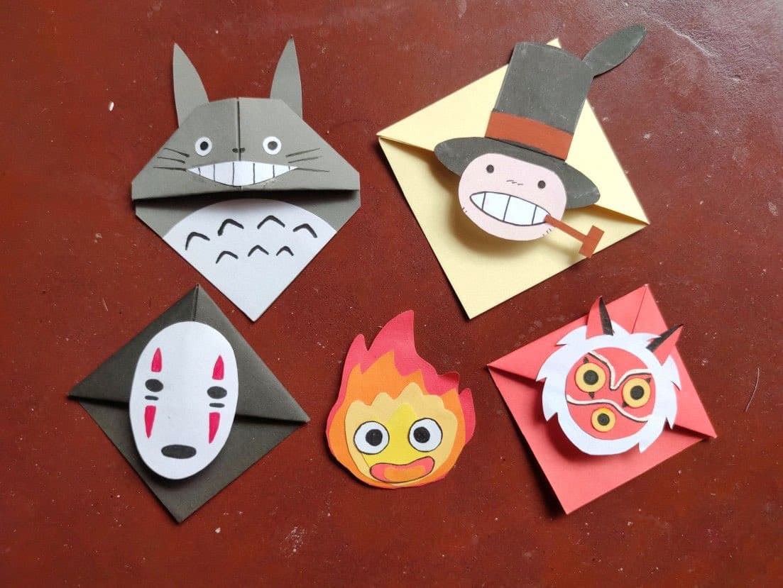 A photograph showing five handmade paper crafts on a red surface, depicting characters from Studio Ghibli films: Totoro, Totoro with a hat, No-Face, a fire spirit, and a red mask.