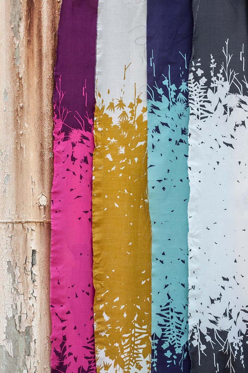 A close-up photograph of five vertical strips of fabric hanging side-by-side against a weathered, peeling wooden surface. Each strip features a different colorway of a botanical print with silhouetted flowers and foliage, including magenta, gold, teal, and white on dark backgrounds.