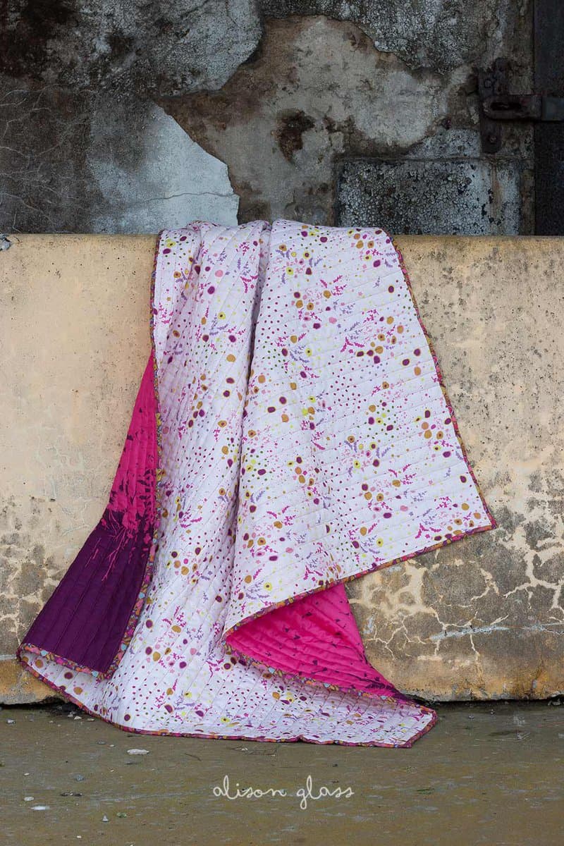 A quilted textile with a white background and a colorful, abstract dot pattern is draped over a weathered concrete wall, revealing a bright pink and purple backing. The fabric features visible stitching lines and a decorative border, set against a rustic, textured background.