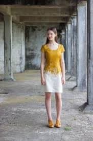 A young woman stands in a dilapidated, concrete corridor wearing a short-sleeved dress with a yellow top and white bottom, paired with yellow shoes.