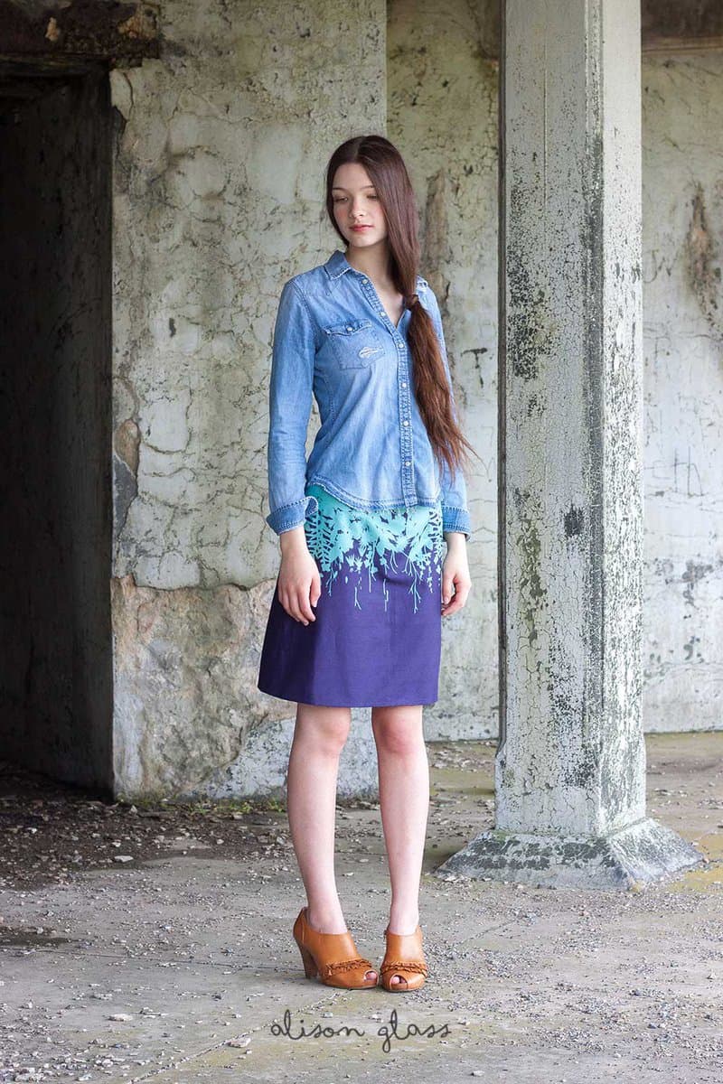 A full-length fashion photograph of a woman standing in a weathered, concrete structure, wearing a denim shirt, a patterned blue and purple skirt, and brown wedge sandals. A watermark reading 'alison glass' is visible at the bottom of the image.