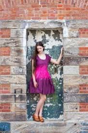 A woman stands in a stone doorway of a brick building, wearing a purple dress with a pink sash and brown shoes. She is posing with one hand on the doorframe.