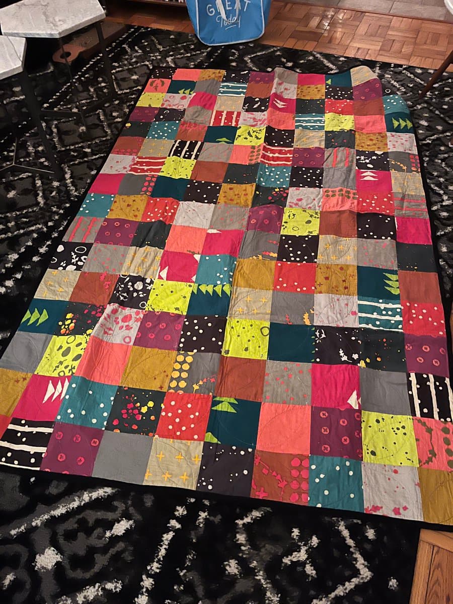 A colorful patchwork quilt made of many square fabric blocks is spread out on a black and white patterned rug. A blue tote bag with the text 'GREAT' is visible at the top of the frame.