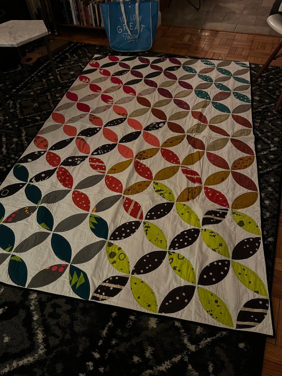 A colorful patchwork quilt featuring a repeating leaf pattern in shades of red, green, blue, and black is spread out on a dark patterned rug. In the background, a blue tote bag with white text sits on a wooden floor near a bookshelf.