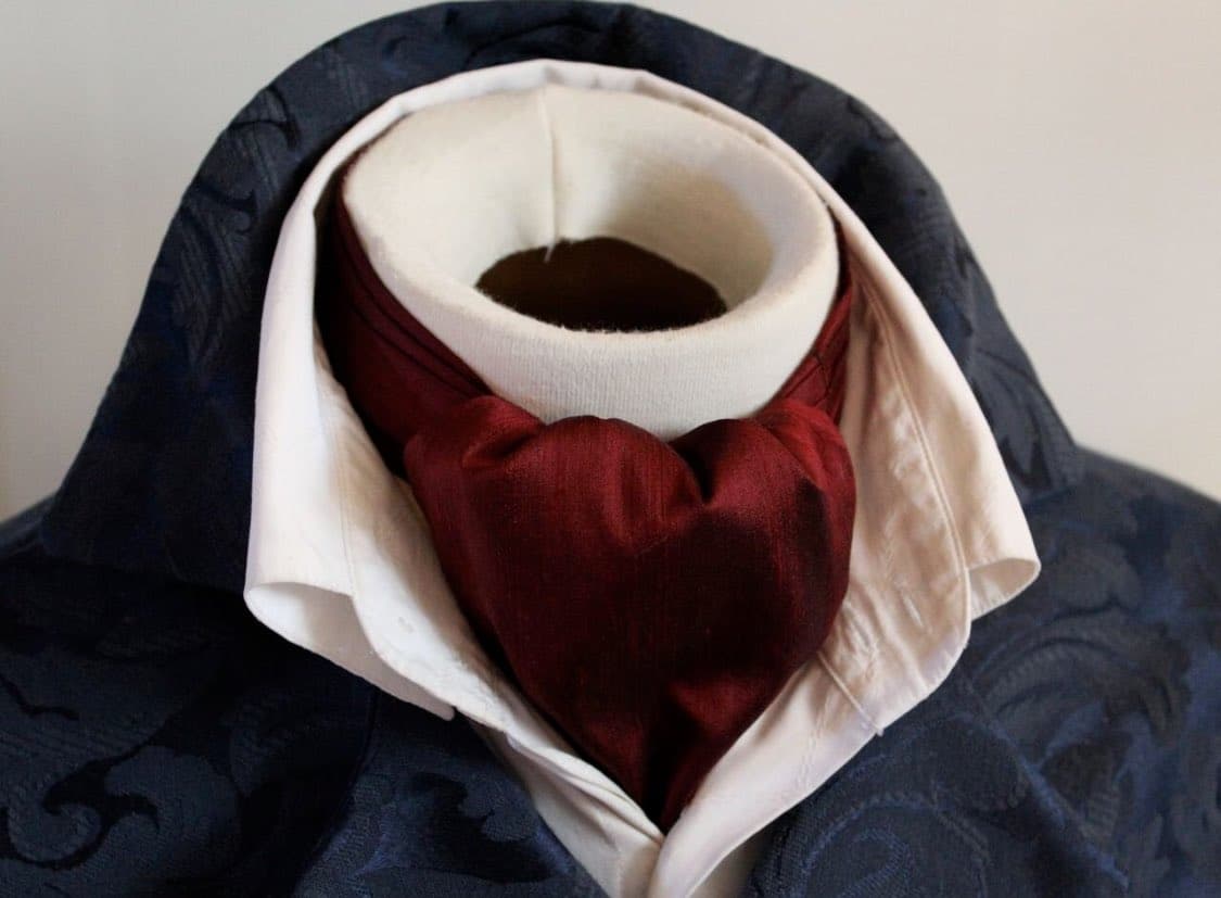 A close-up product photograph of a mannequin neck wearing a dark blue patterned jacket, a white shirt collar, and a maroon cravat.