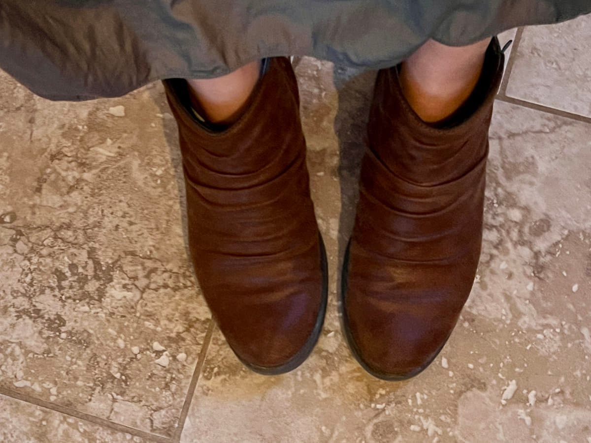 A top-down view of a person's feet wearing brown, slouchy leather ankle boots on a light-colored tiled floor.