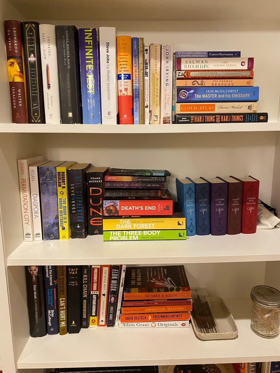 A photo of a white bookshelf filled with numerous books, including titles by Leonardo da Vinci, Salman Rushdie, Iain M. Banks, and J.R.R. Tolkien. The books are arranged both vertically and horizontally across three visible shelves, with a small dish and a glass jar on the bottom right.