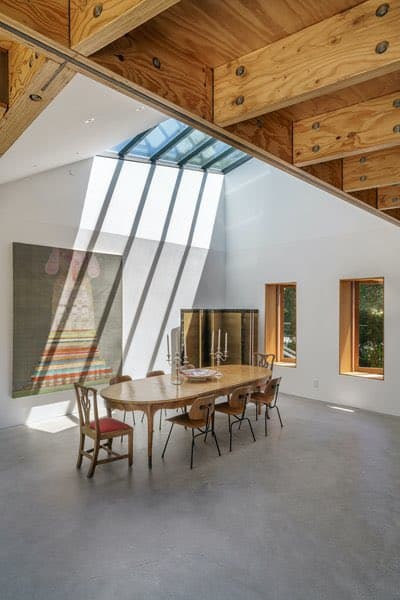 A photograph of a modern dining room featuring a long wooden table and chairs beneath a large, angled skylight that casts diagonal shadows on the white wall. The room has exposed wooden ceiling beams, a concrete floor, and a large abstract painting on the left wall.