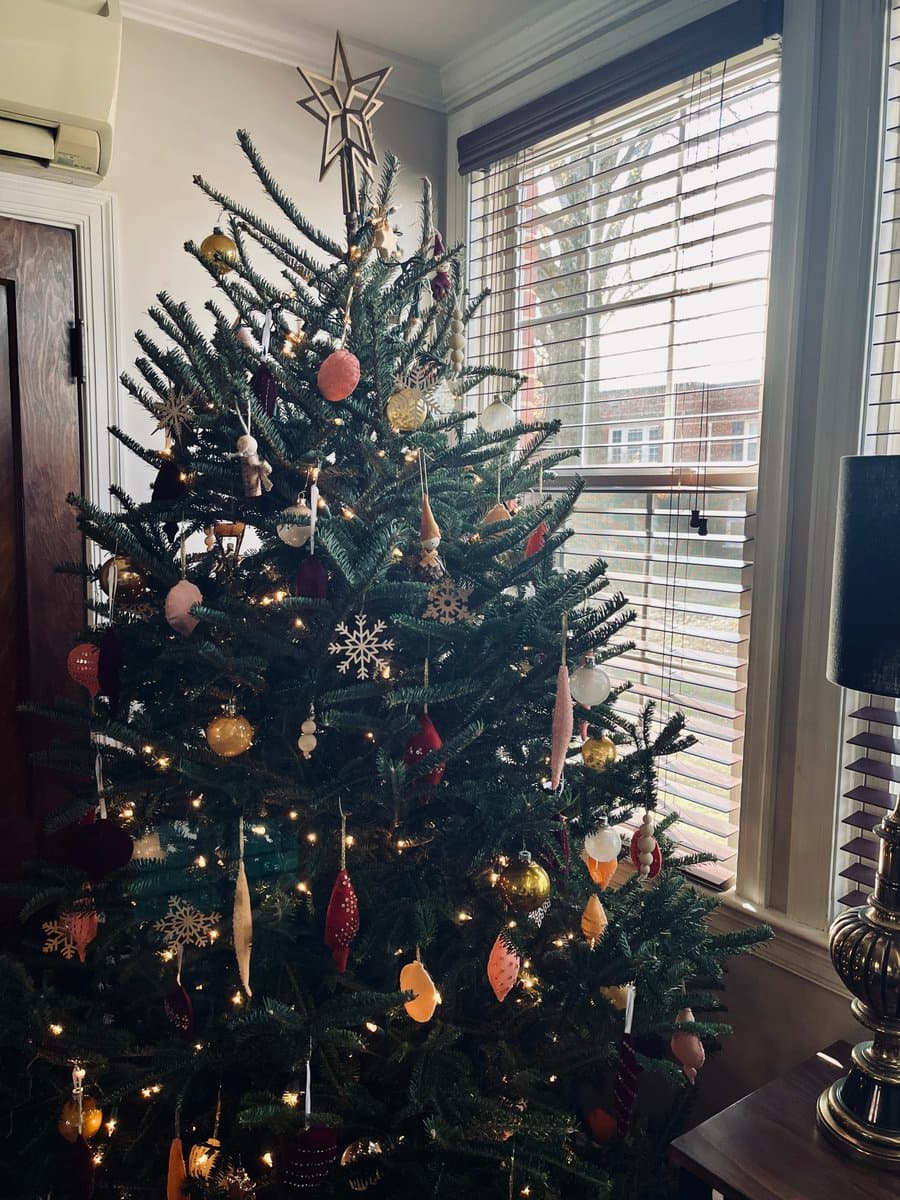 A decorated Christmas tree stands in a corner of a room, adorned with gold, red, and white ornaments and topped with a star. The tree is positioned next to a window with white blinds, with a lamp visible on a side table to the right.