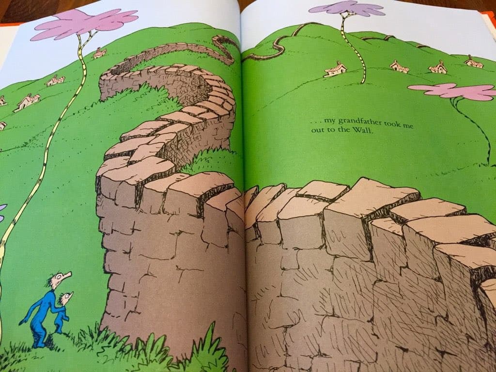 A photograph of an open picture book featuring a cartoon illustration of a stone wall winding across green hills, with two small figures in blue standing at the base of the wall on the left page.