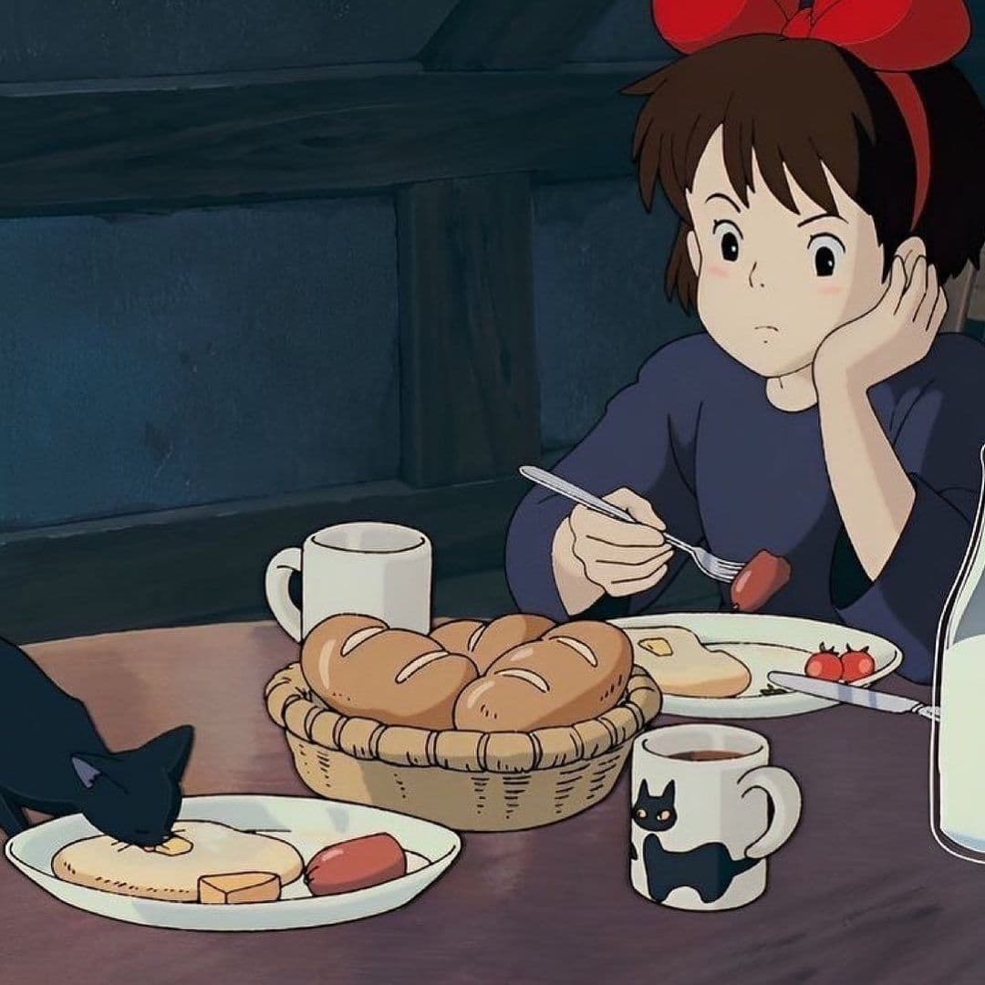 A still from the animated film 'Kiki's Delivery Service' showing a young girl with a red bow sitting at a table with a breakfast spread, including bread, eggs, and a black cat eating from a plate.
