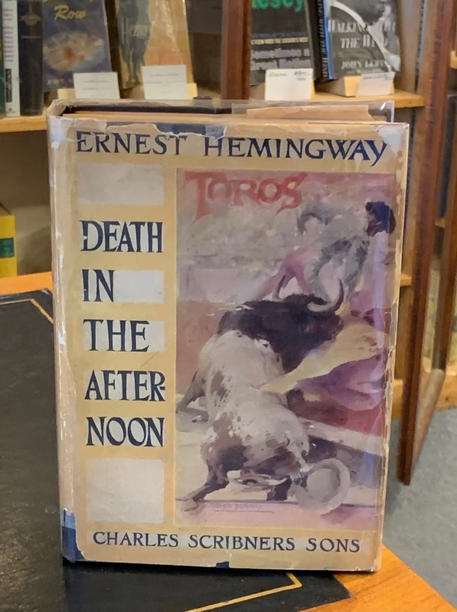 A photograph of a vintage, first-edition copy of Ernest Hemingway's book 'Death in the Afternoon' standing on a wooden surface in a bookstore. The book's cover features a painted illustration of a bullfighting scene and is wrapped in protective plastic.