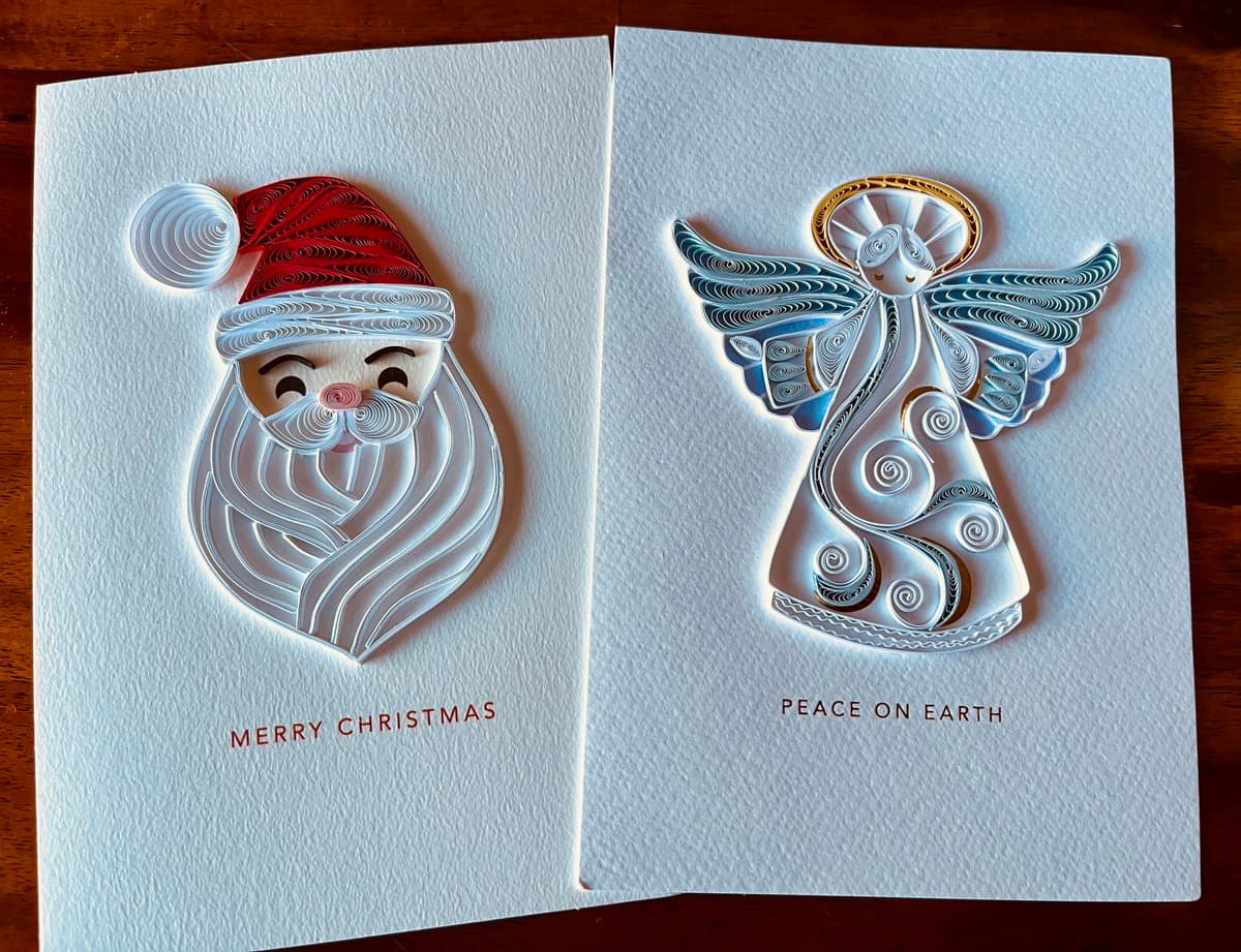 A photograph of two white Christmas cards featuring intricate quilling art. The left card displays a portrait of Santa Claus, while the right card features an angel with a halo and wings.