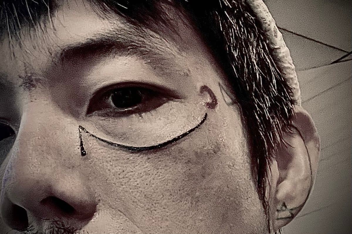 A close-up, black-and-white photograph of a person's face featuring face paint, including a line under the eye ending in a teardrop shape and a small symbol on the temple.