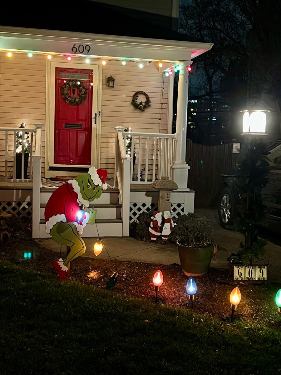 A nighttime photo of a house's front porch decorated with Christmas lights and a large inflatable Grinch figure wearing a Santa hat. The Grinch is positioned on the lawn, holding a string of lights, with the house number 609 visible on the porch and a matching number sign on the ground.